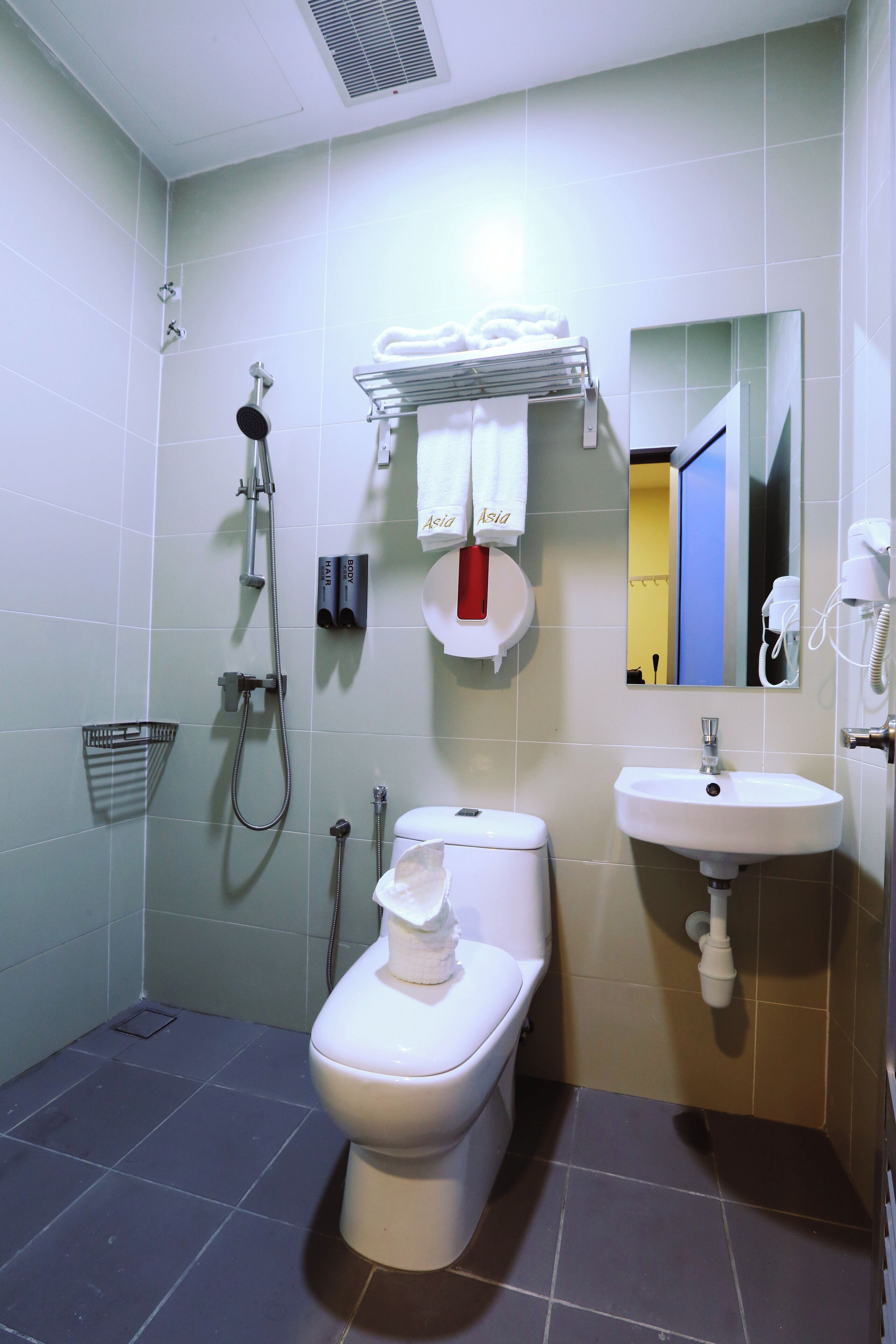 A bathroom with a toilet, sink, and shower. There is a towel rack with towels, a mirror, and a trash bin.