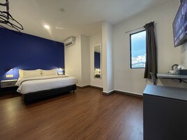 A bedroom with a bed, bedside lamps, a window with curtains, an air conditioning unit, and a view of buildings outside.