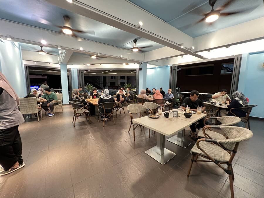 Un comedor amplio con techos altos, ventiladores de techo e iluminación abundante. Las mesas están dispuestas para grupos, y el piso está cubierto de baldosas.