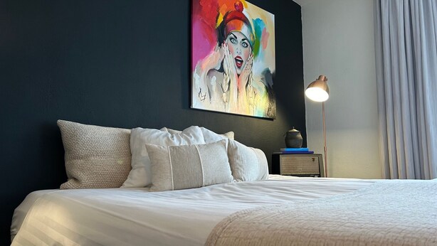 A neatly made bed with white linens and multiple pillows, a bedside table with a lamp, and a colorful abstract painting on the wall. - Hotel BE41 (Maastricht)