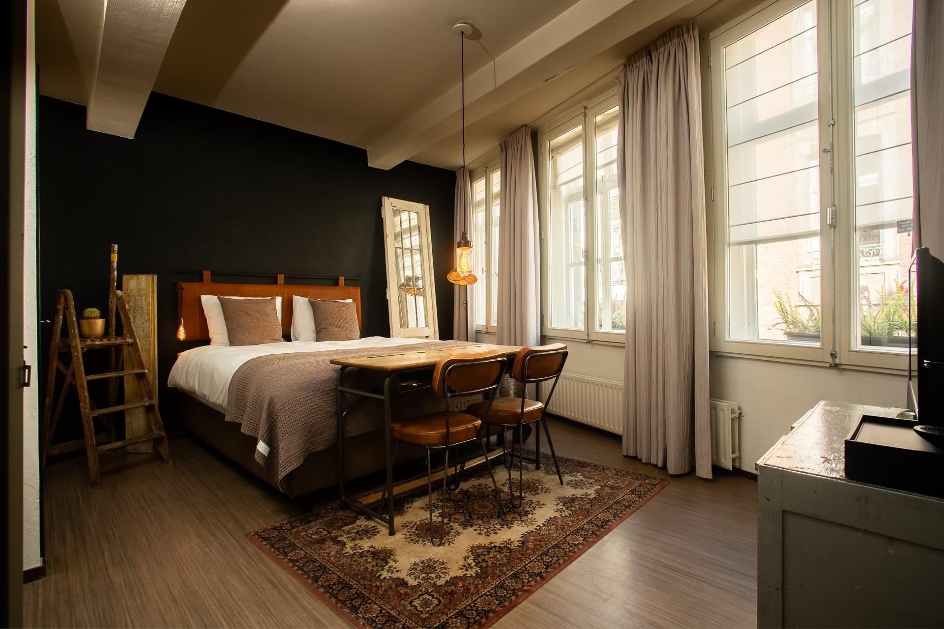 A bedroom with a bed, a desk, and a chair. There is a window with curtains, a radiator, and a lamp.
