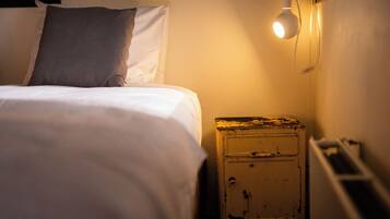 Een bed met witte lakens en één kussen, een klein nachtkastje met een lamp, en een wandlamp.