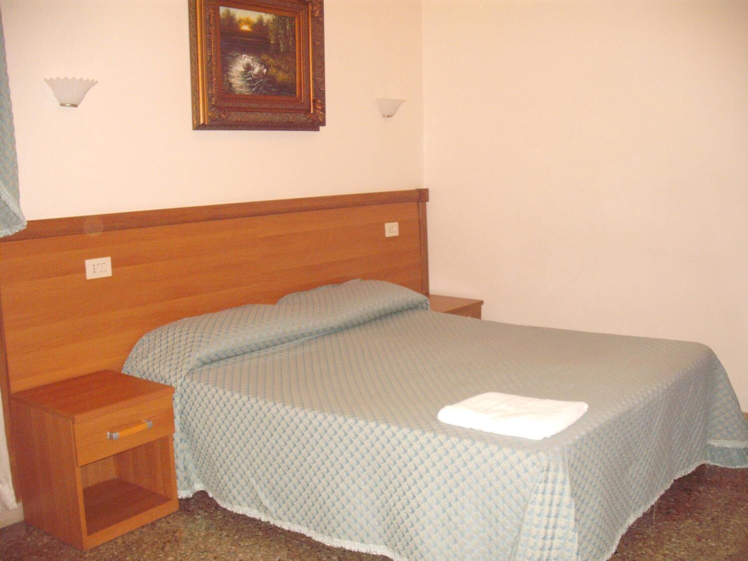 A simple bedroom with a wooden headboard, a single bed with a blue quilt, a nightstand, and a framed picture on the wall.