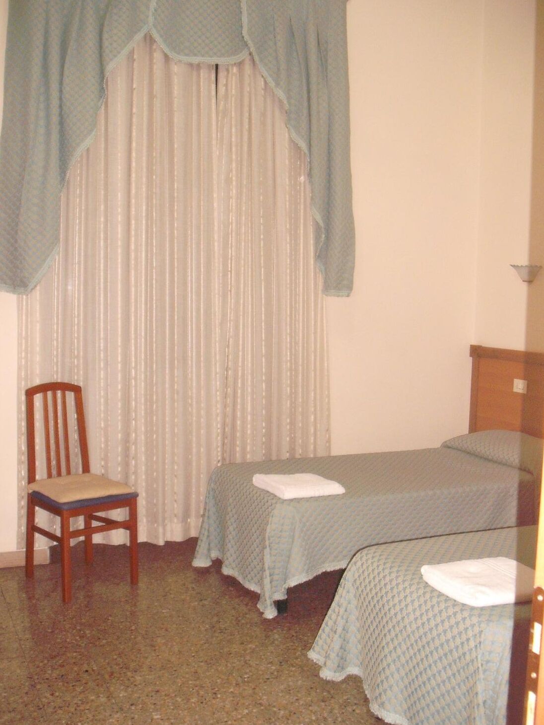A small, simple room with two single beds, a wooden chair, and a window with sheer curtains.