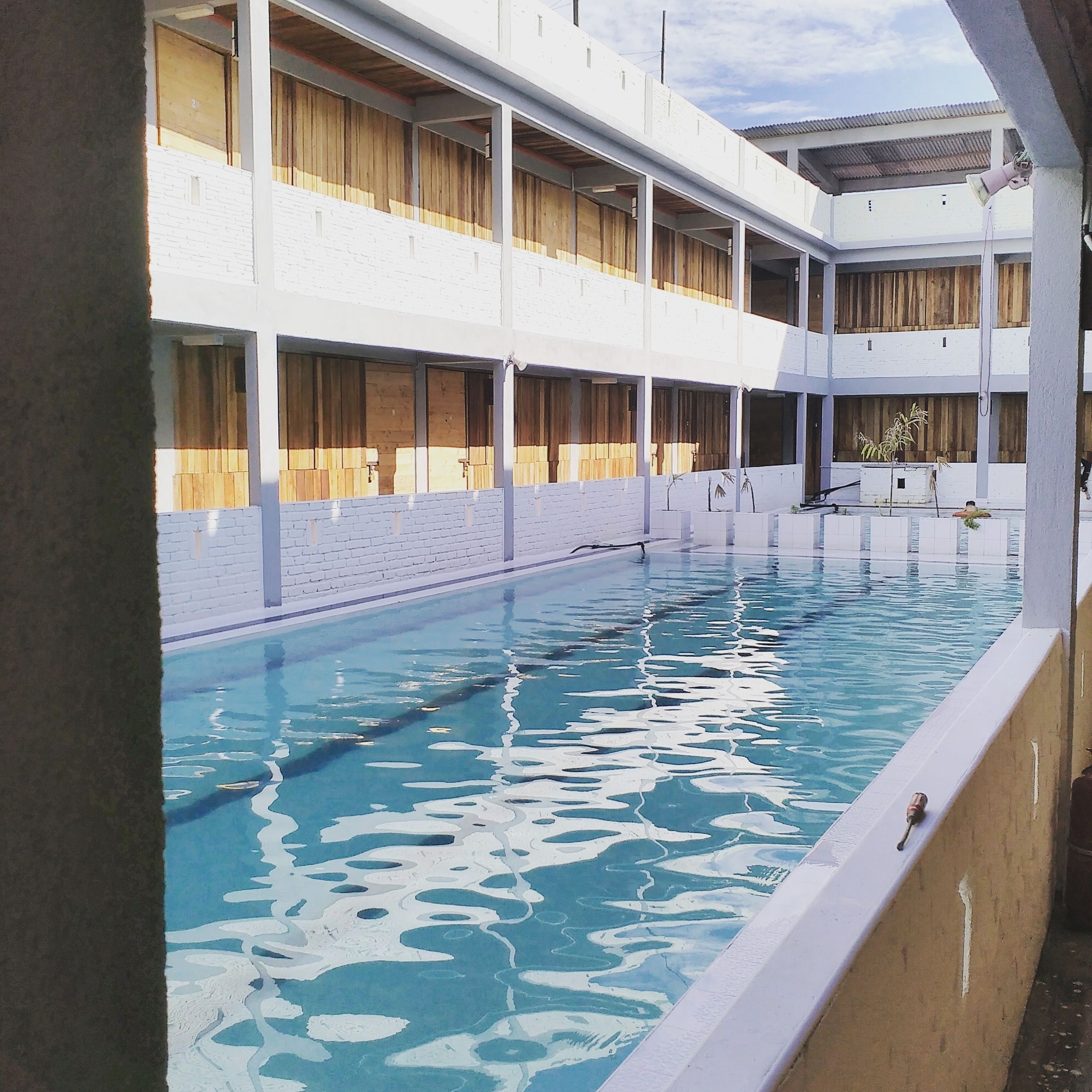 A swimming pool with a white poolside area, wooden deck, and a multi-level building with large windows.