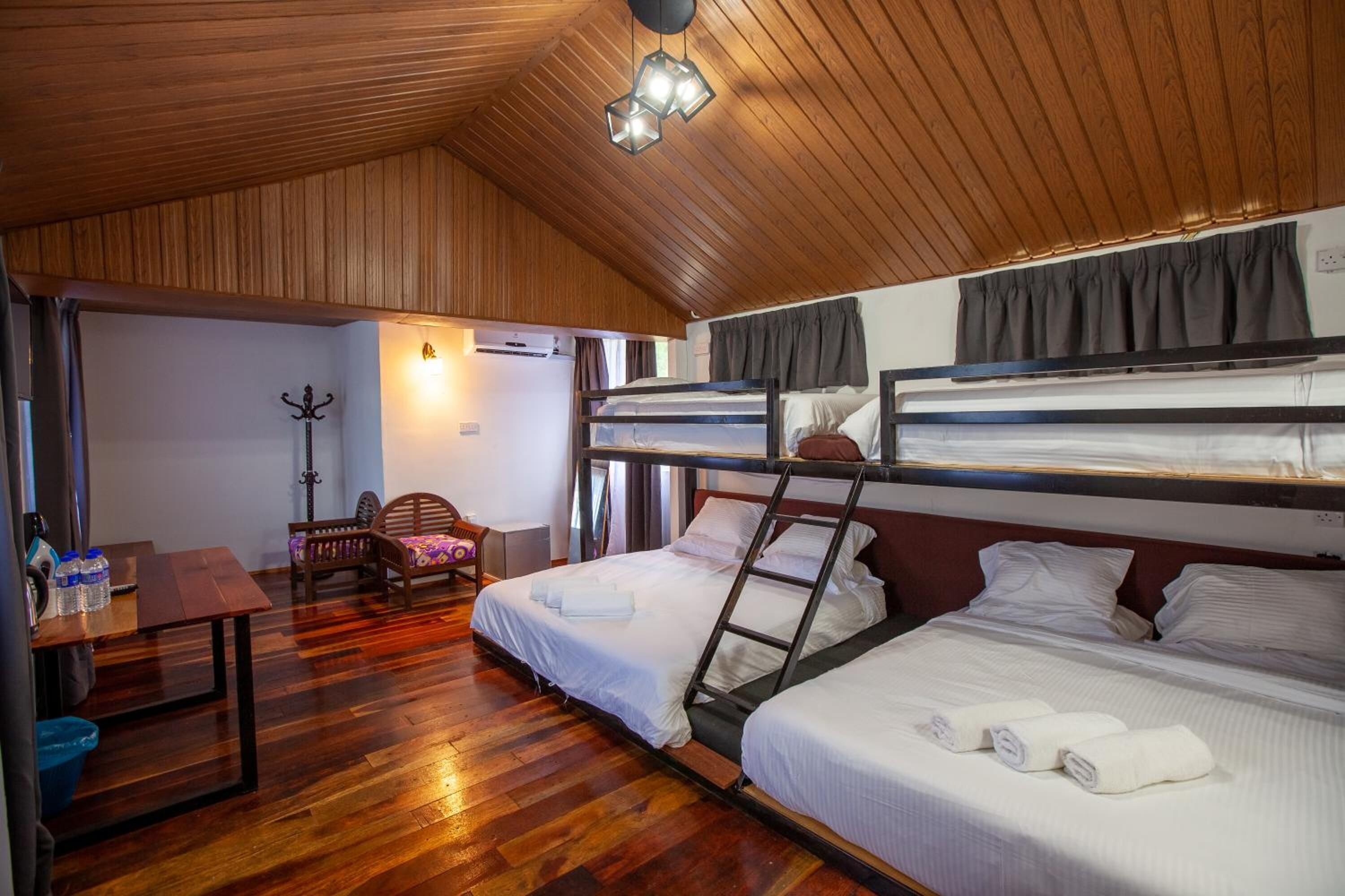 Habitación pequeña con literas, techo de madera, escritorio con silla y una mesita con botellas de agua.