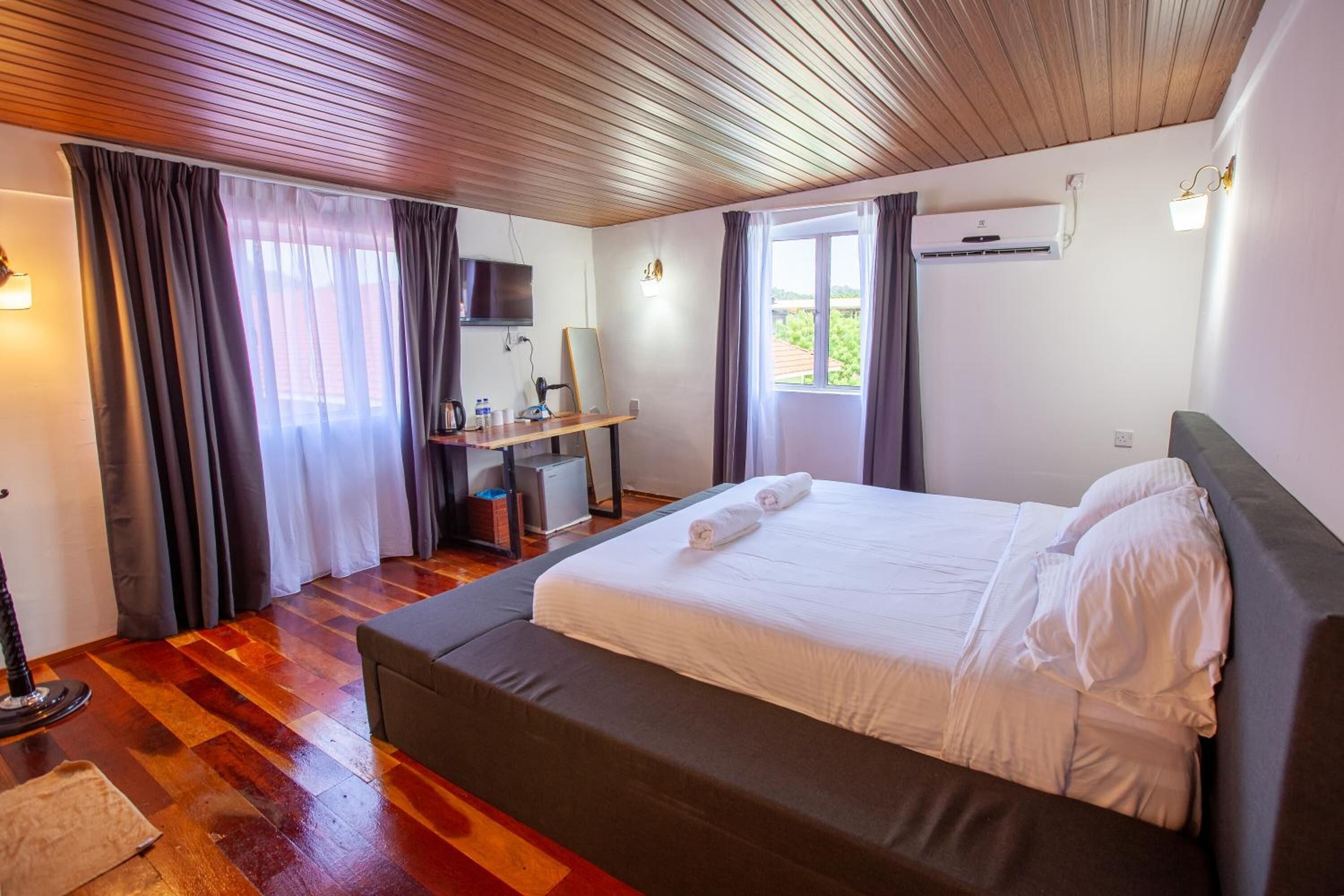 A hotel room with a wooden ceiling, a bed with white linens, a desk with a computer, and a window with curtains.