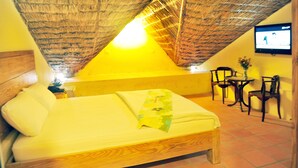 A room with a bed, a small table, two chairs, a TV on the wall, and a thatched ceiling.