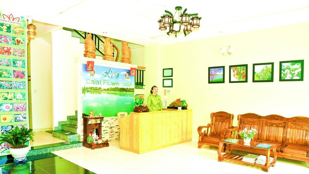 A reception area with a wooden desk, a receptionist, a reception desk with a floral backdrop, a staircase, a bench, and framed pictures on the wall.