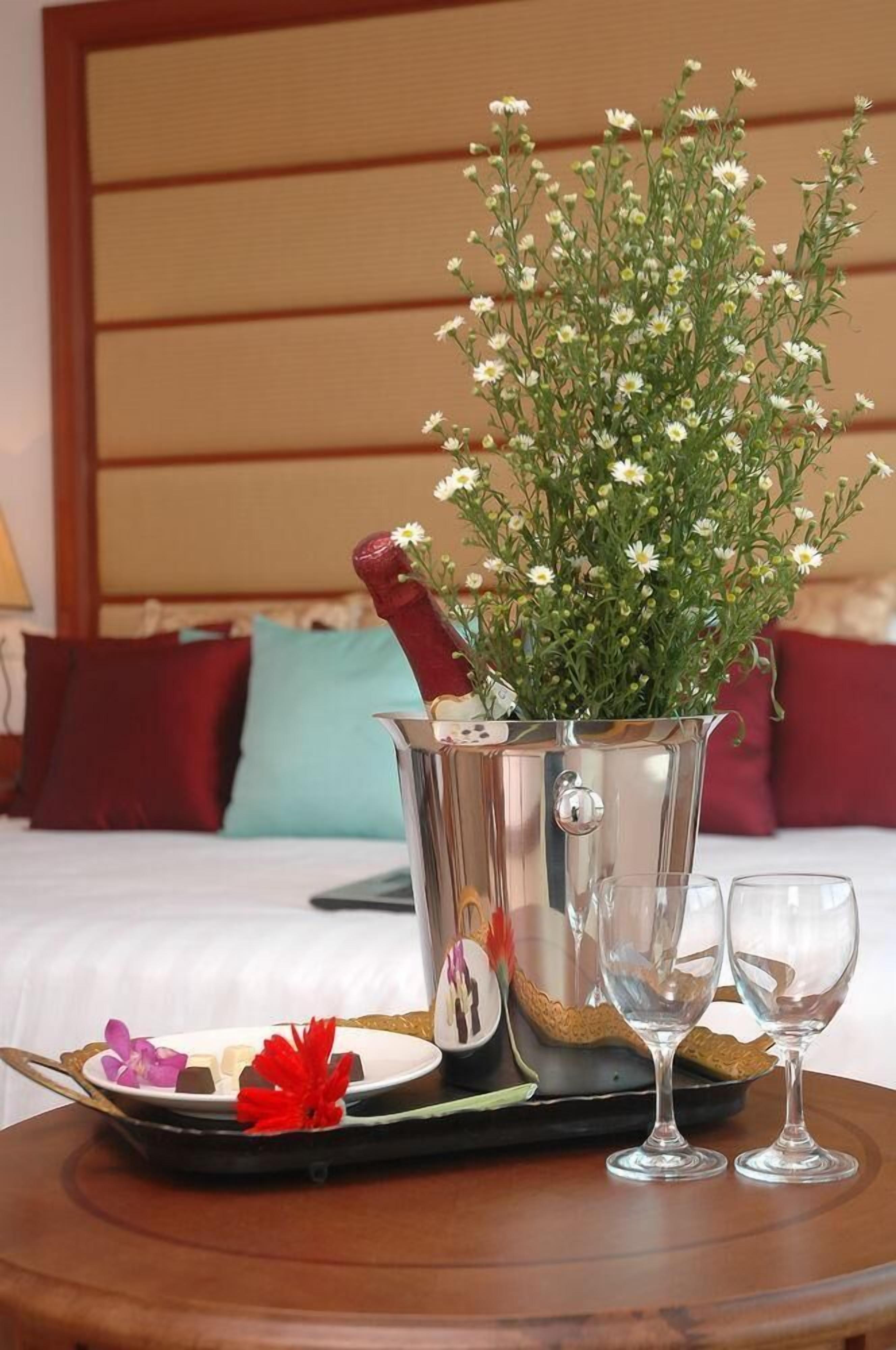A hotel room with a bed, a bedside table with a tray of food and drinks, and a potted plant with white flowers.