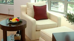 A modern living room with a white sofa, a red pillow, and a round table with a fruit bowl. Large windows offer a city view. - Oriental Crystal Hotel Kajang (Kajang)