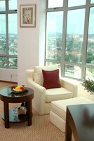 A modern living room with a white sofa, a red pillow, and a round table with a fruit bowl. Large windows offer a city view.