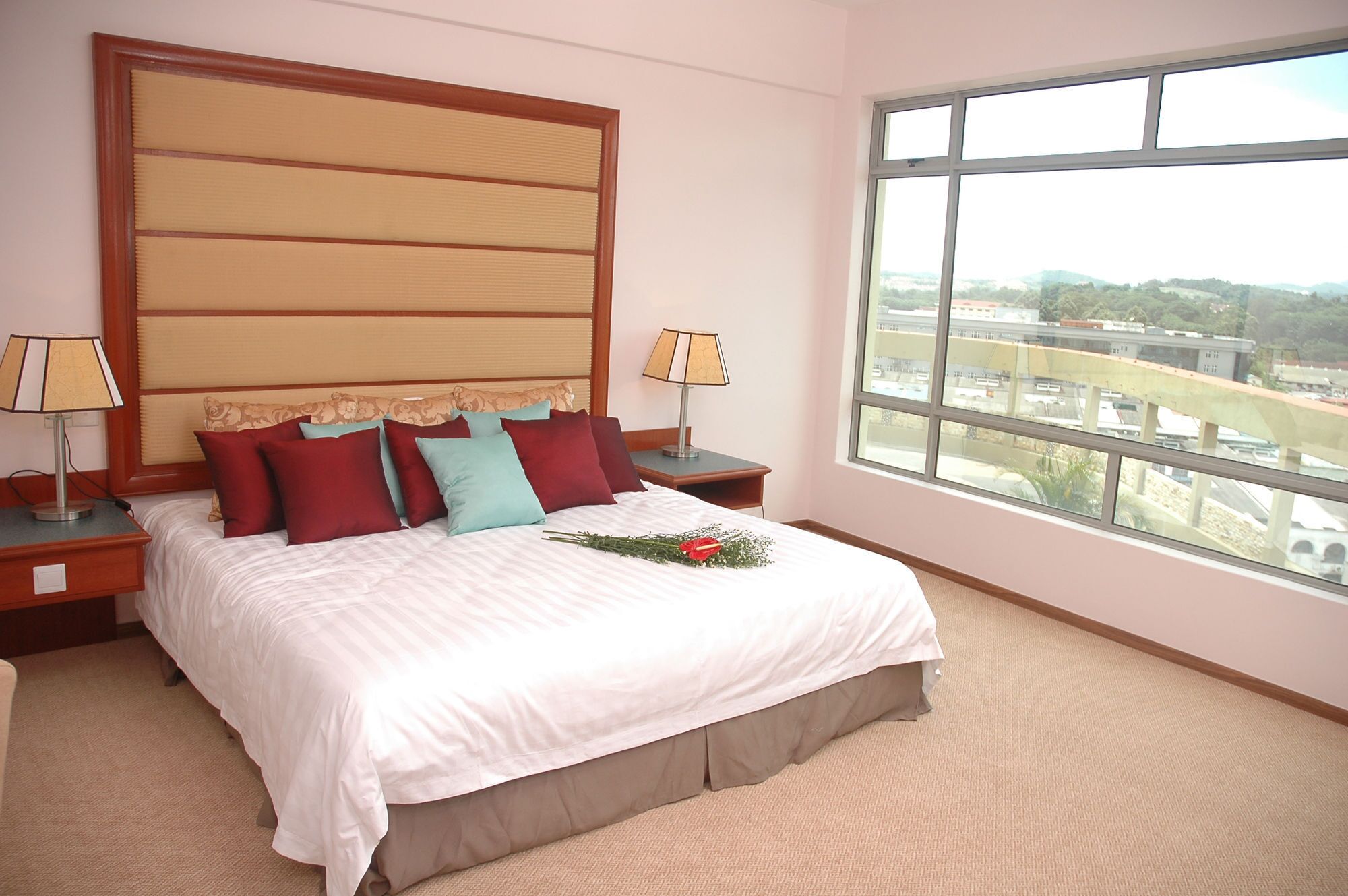 A hotel room with a large bed, two bedside tables with lamps, a view of the city through a large window, and a decorative headboard.