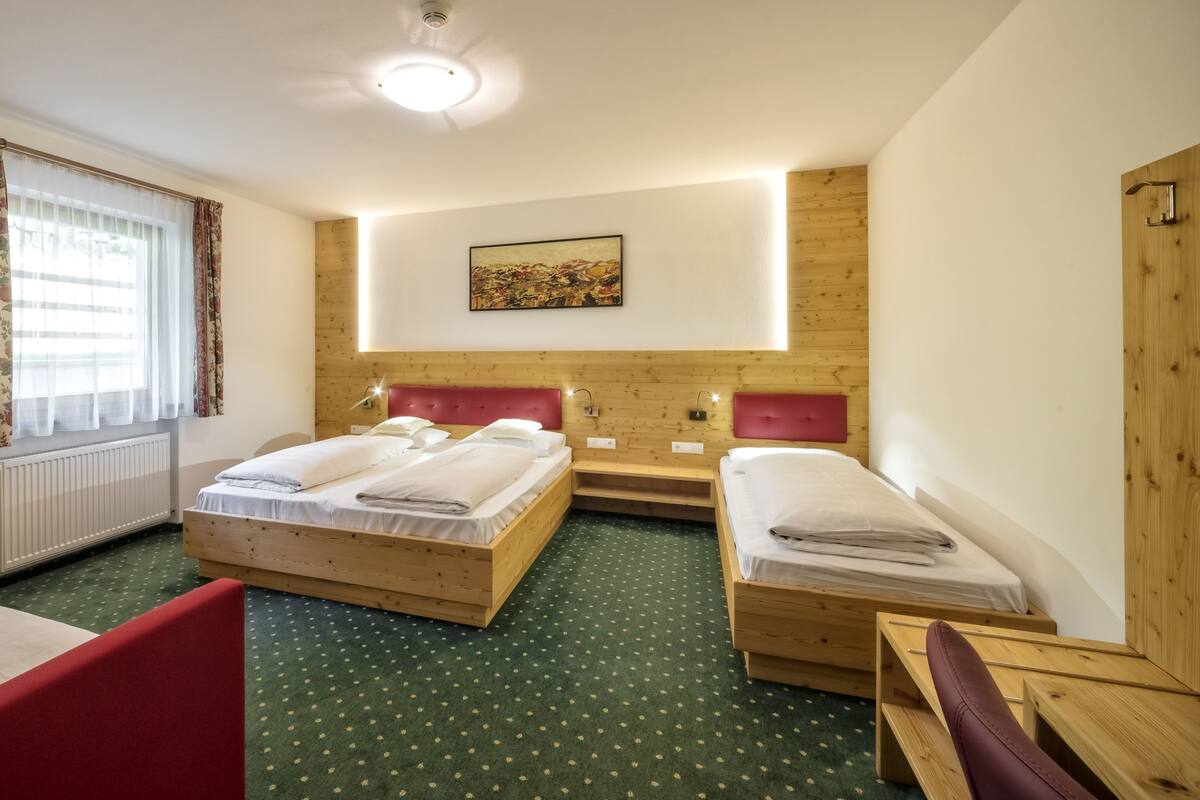 Una habitación de hotel con dos camas de madera, un cabecero rojo, una alfombra verde con lunares blancos, un radiador y una ventana con cortinas estampadas.