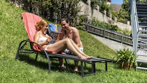 Two people relaxing on a sun lounger in a grassy area with a stone wall and a tower in the background. - Hotel Elisabeth (Chienes)