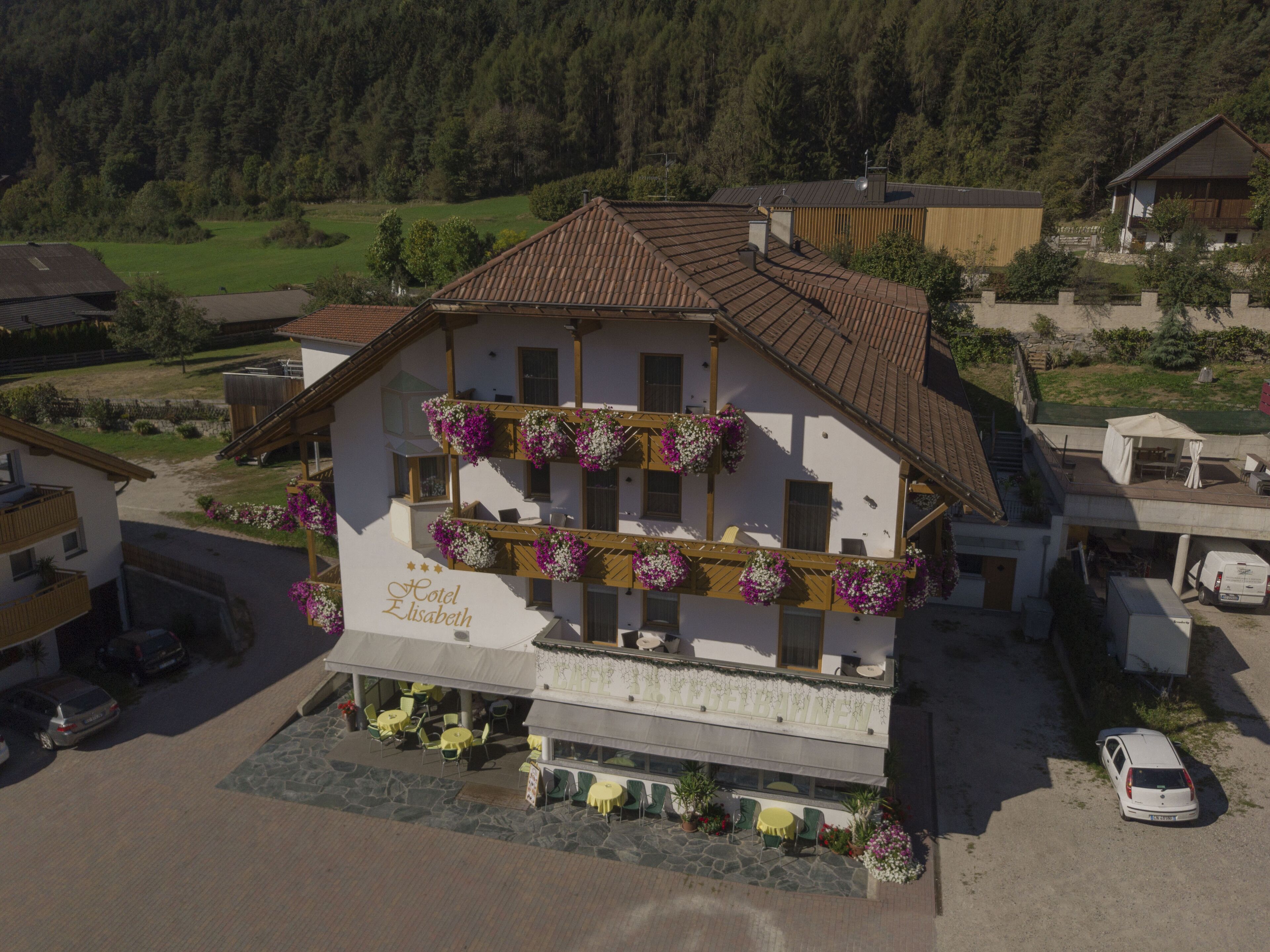 Ein zweistöckiges Gebäude mit Balkonen, die mit Blumen geschmückt sind, einem Schild mit der Aufschrift 'Hotel Elisabeth', Außenbestuhlung und einem Parkplatz.