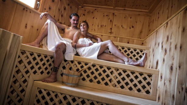 A wooden sauna with two people sitting on wooden benches, a wooden lattice wall, and a wooden bucket. - Hotel Elisabeth (Chienes)