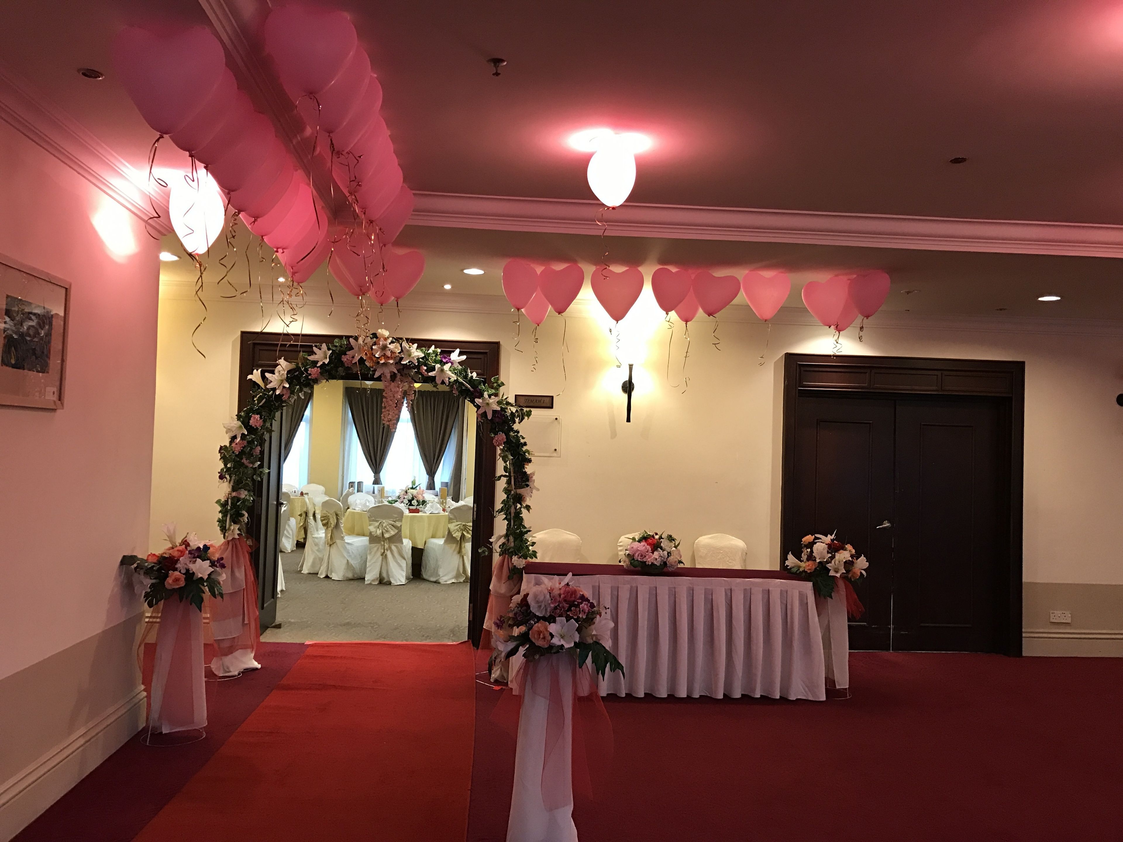 A room with a red carpet, white chairs, a floral archway, and pink balloons.