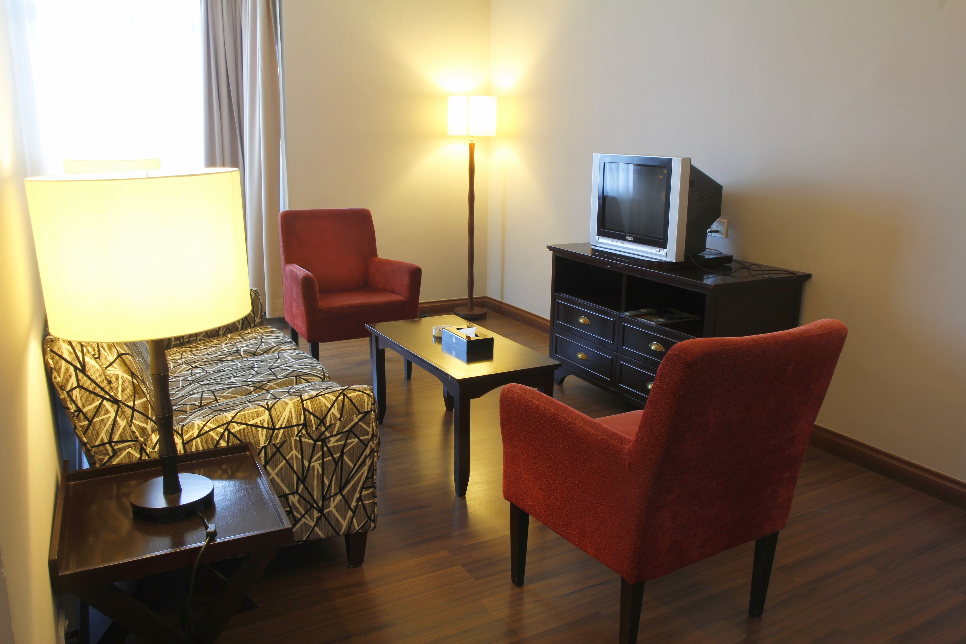 Una habitación de hotel con una cama, un sillón rojo, una mesita, un televisor sobre un soporte y una lámpara de pie.