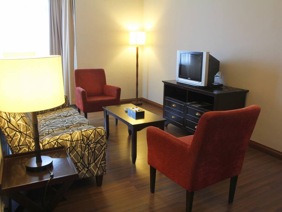 Una habitación de hotel con una cama, un sillón rojo, una mesita, un televisor sobre un soporte y una lámpara de pie.