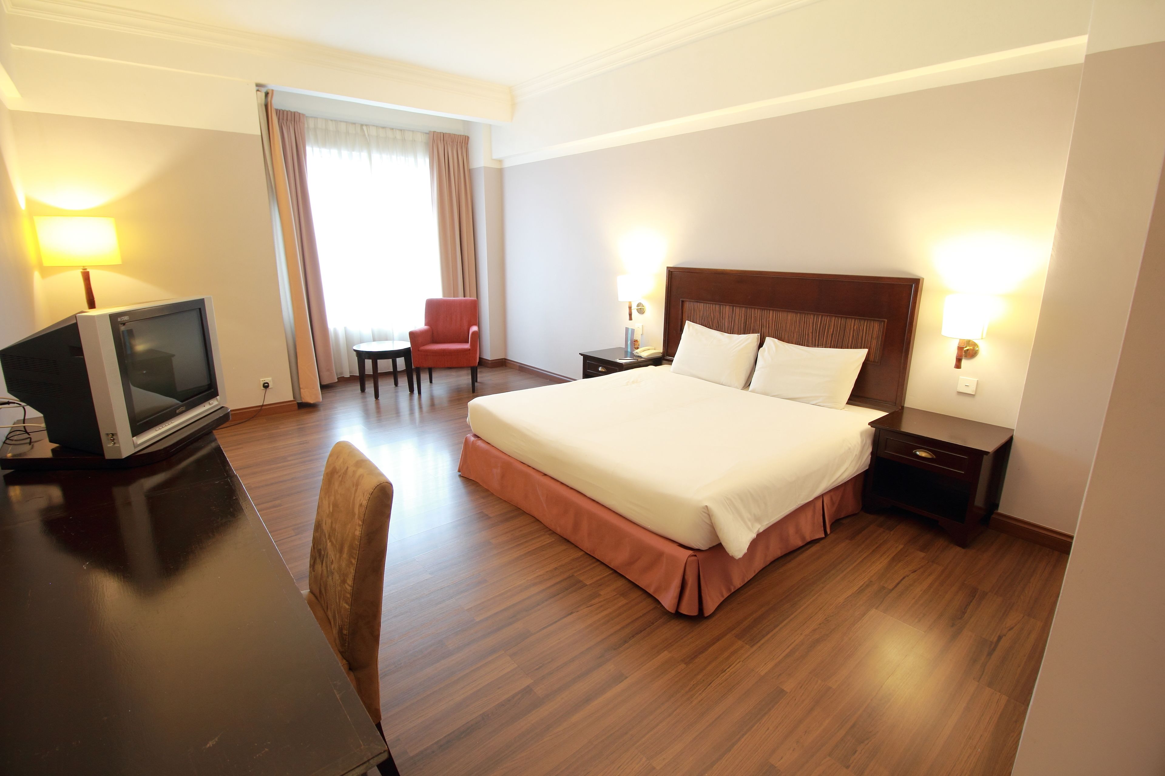 Habitación de hotel con una cama grande, un escritorio con televisor, una silla y dos lámparas de noche.