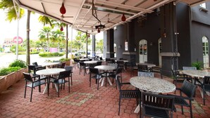 Un área de café al aire libre con mesas y sillas, techo cubierto y vista a palmeras y edificios en el fondo.