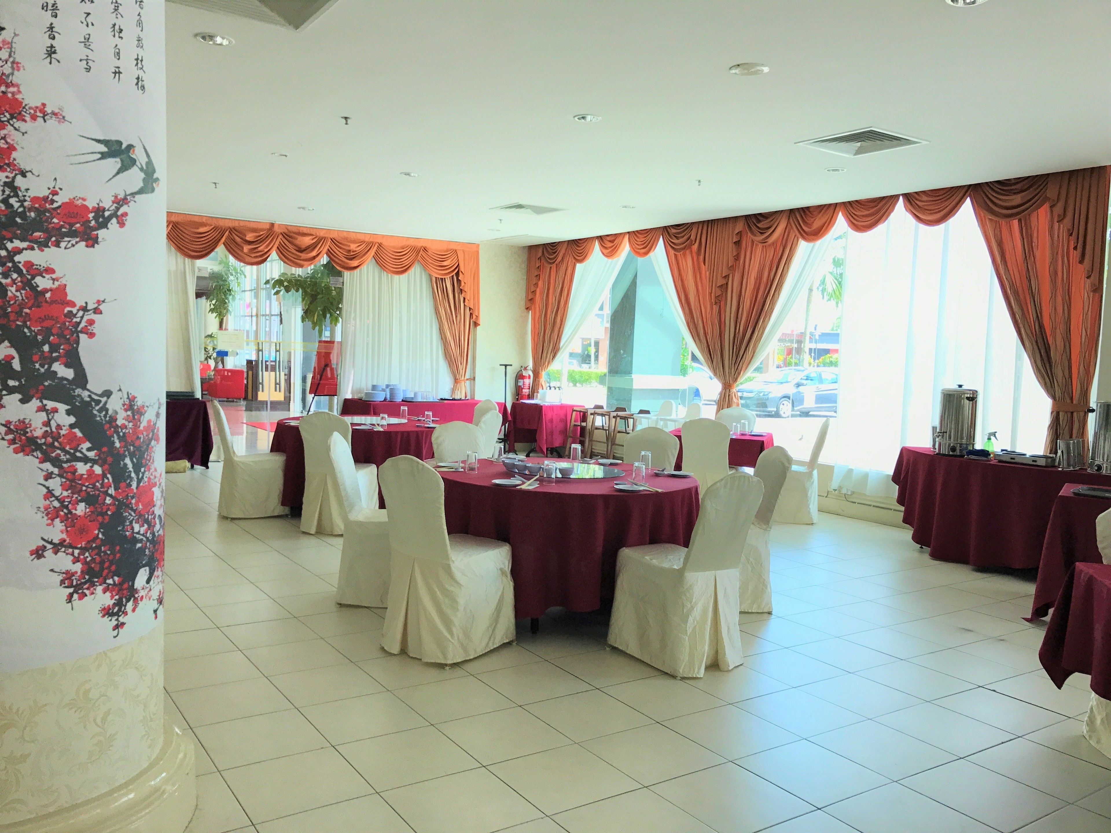 Une salle de banquet avec des tables rondes recouvertes de nappes rouges et des chaises blanches, de grandes fenêtres ornées de rideaux orange et un mur décoratif aux motifs floraux.