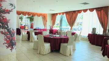 Une salle de banquet avec des tables rondes recouvertes de nappes rouges et des chaises blanches, de grandes fenêtres ornées de rideaux orange et un mur décoratif aux motifs floraux.