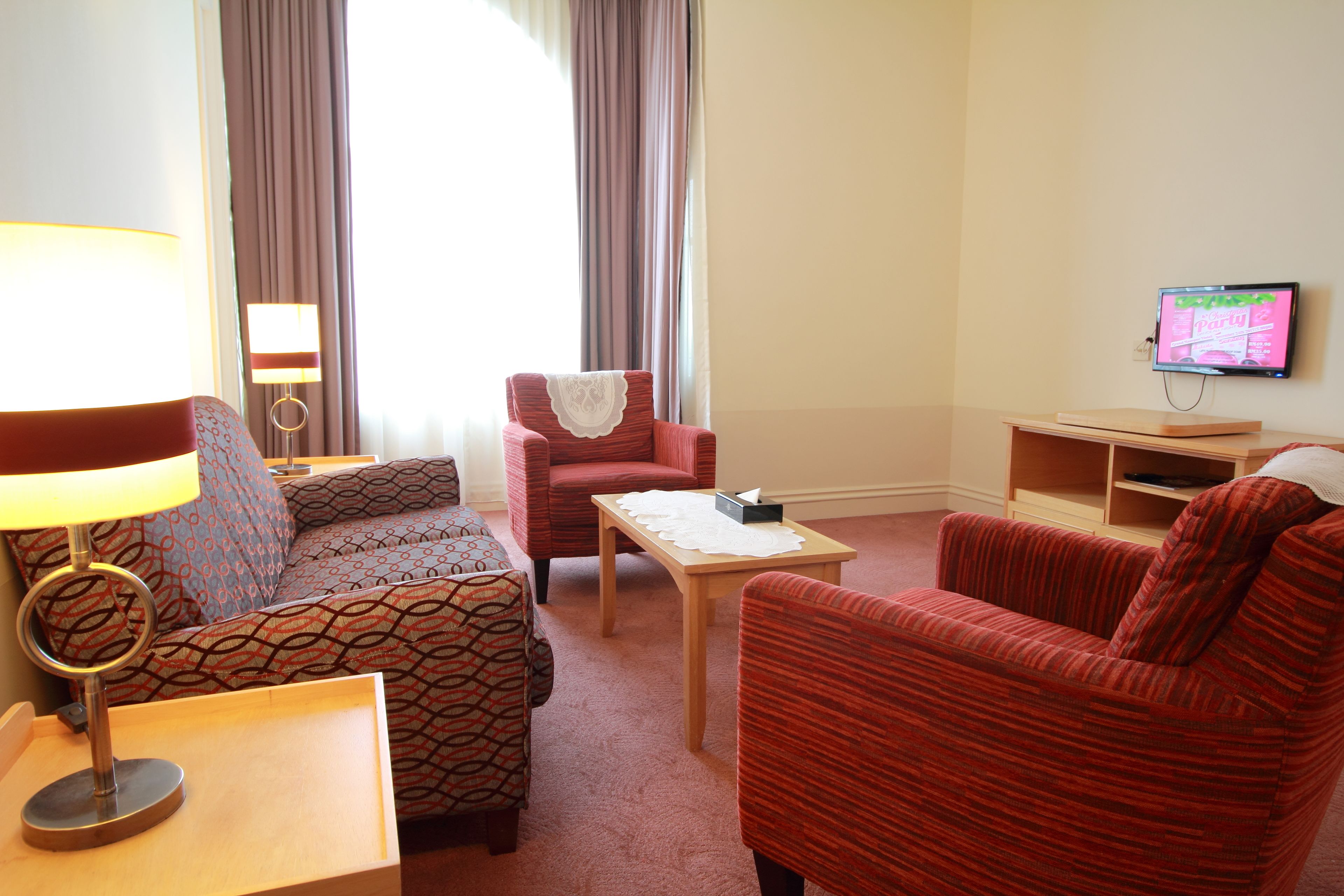 A hotel room with a sofa, armchair, table, lamp, TV, and curtains.