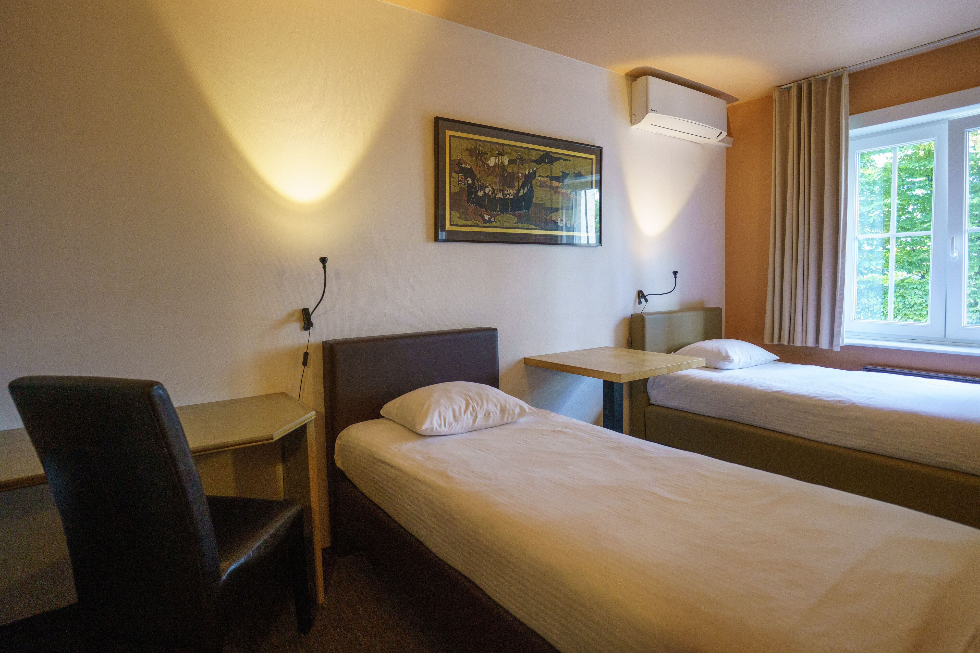 Ett hotellrum med två separata sängar, ett skrivbord, en stol och en tavla på väggen.