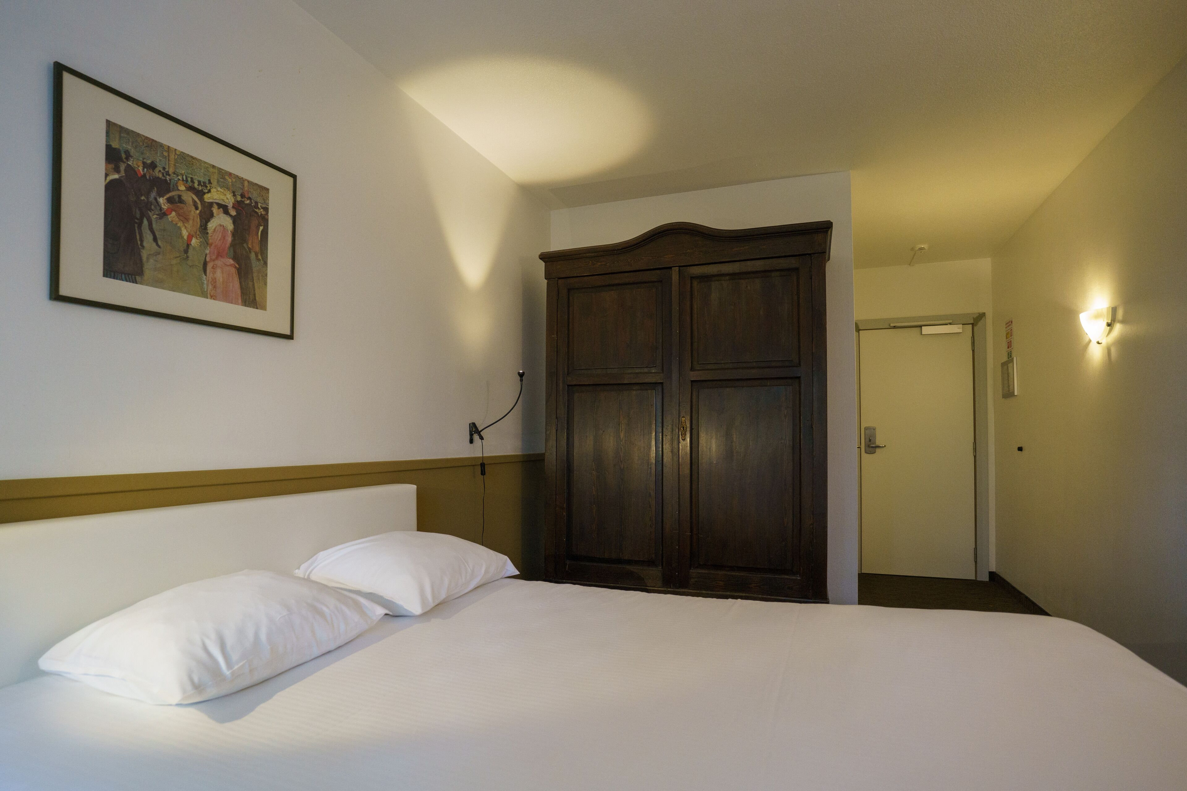 A hotel room with a bed, a large wardrobe, a framed picture on the wall, and a light fixture on the wall.