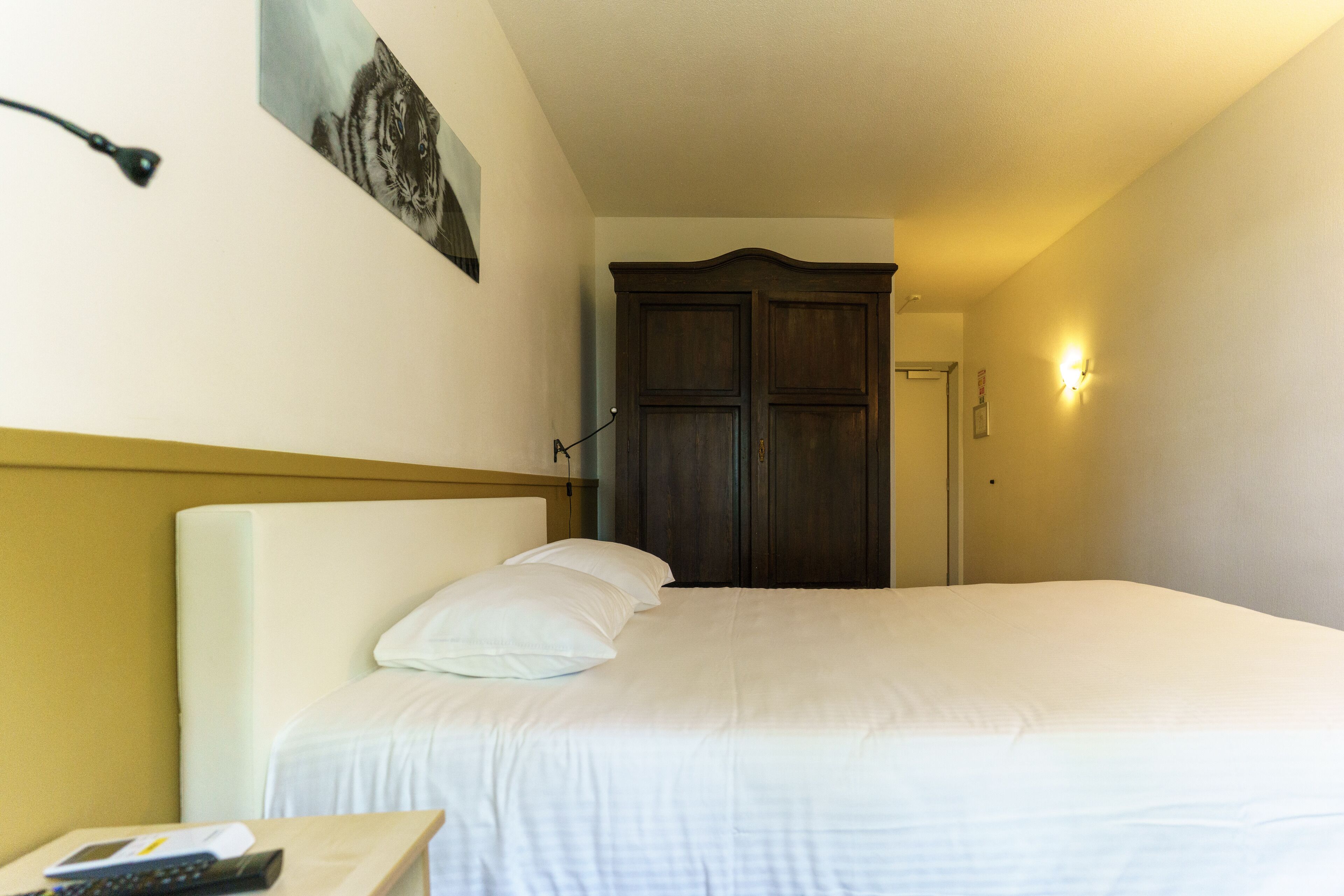 A hotel room with a large bed, a nightstand with a remote control, a wall-mounted lamp, and a door leading to another room.