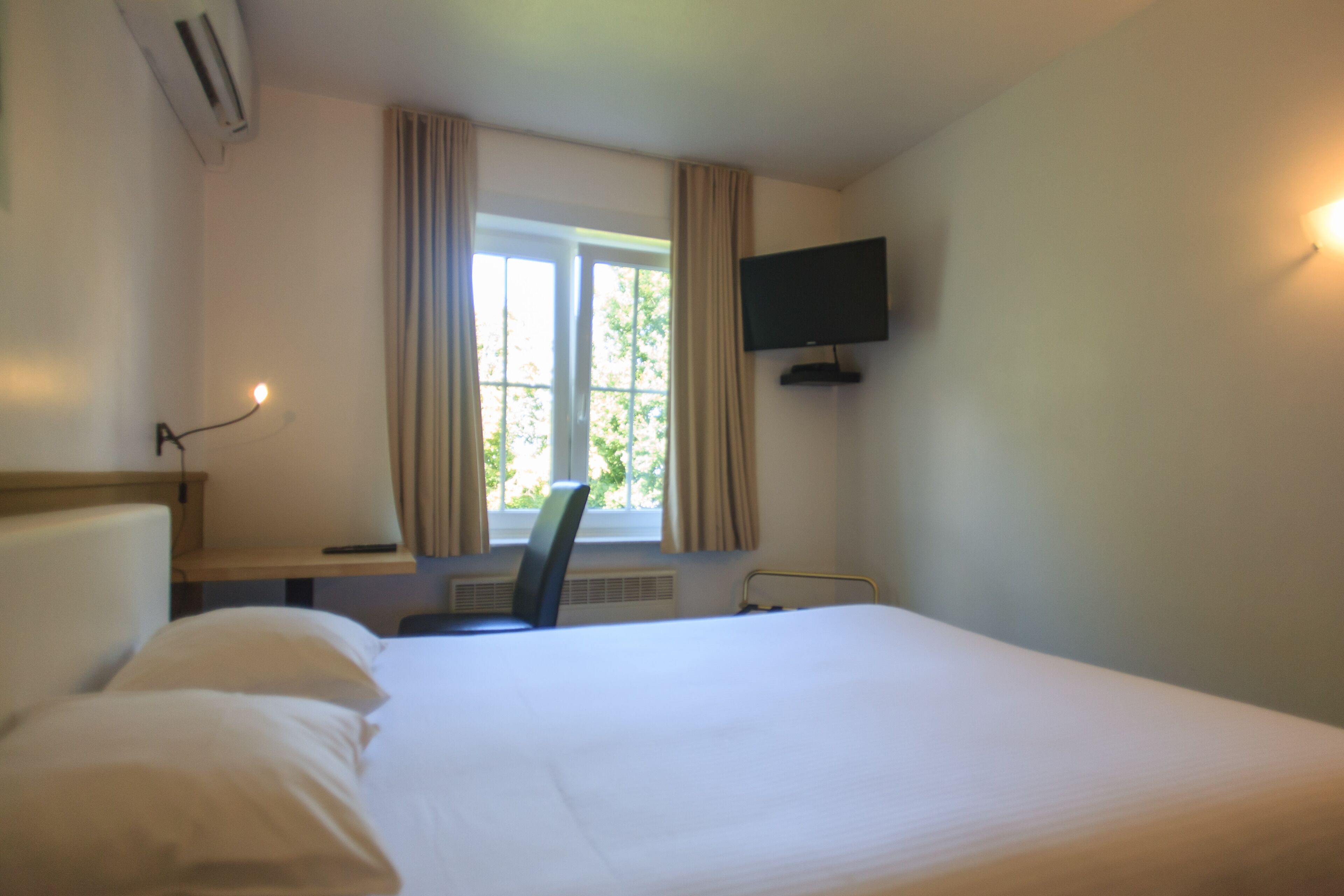 Habitación de hotel con cama, un escritorio con silla, televisor montado en la pared y una ventana con cortinas.