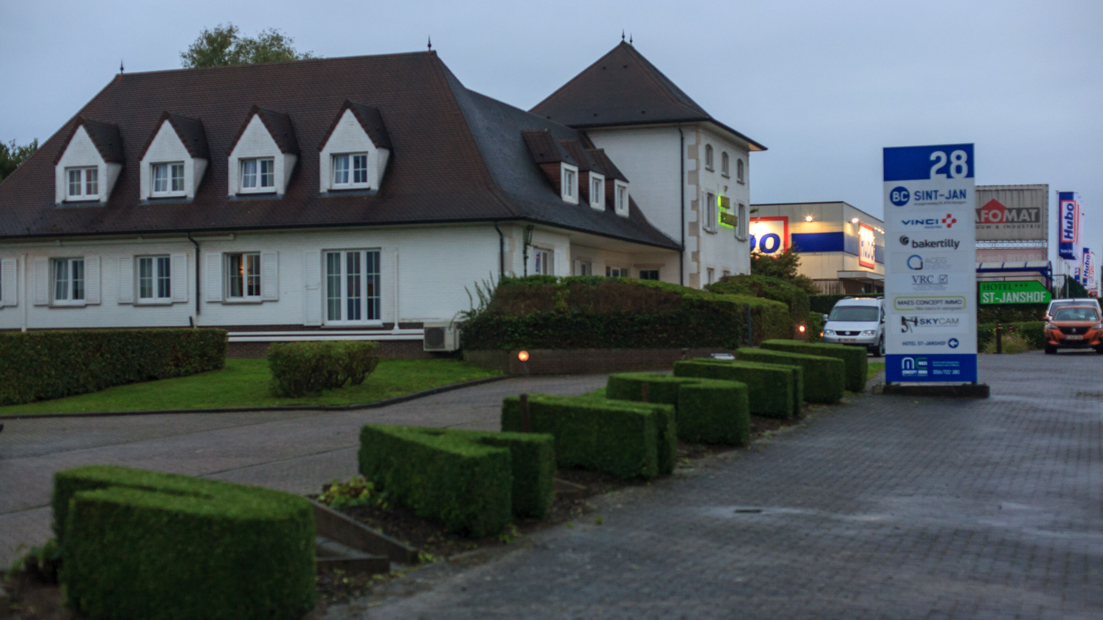 A large, white building with a steeply pitched roof and multiple windows. There is a well-maintained hedge and a paved driveway leading to the entrance.