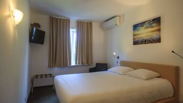 A hotel room with a bed, a television, a window with curtains, an air conditioning unit, and a painting on the wall.