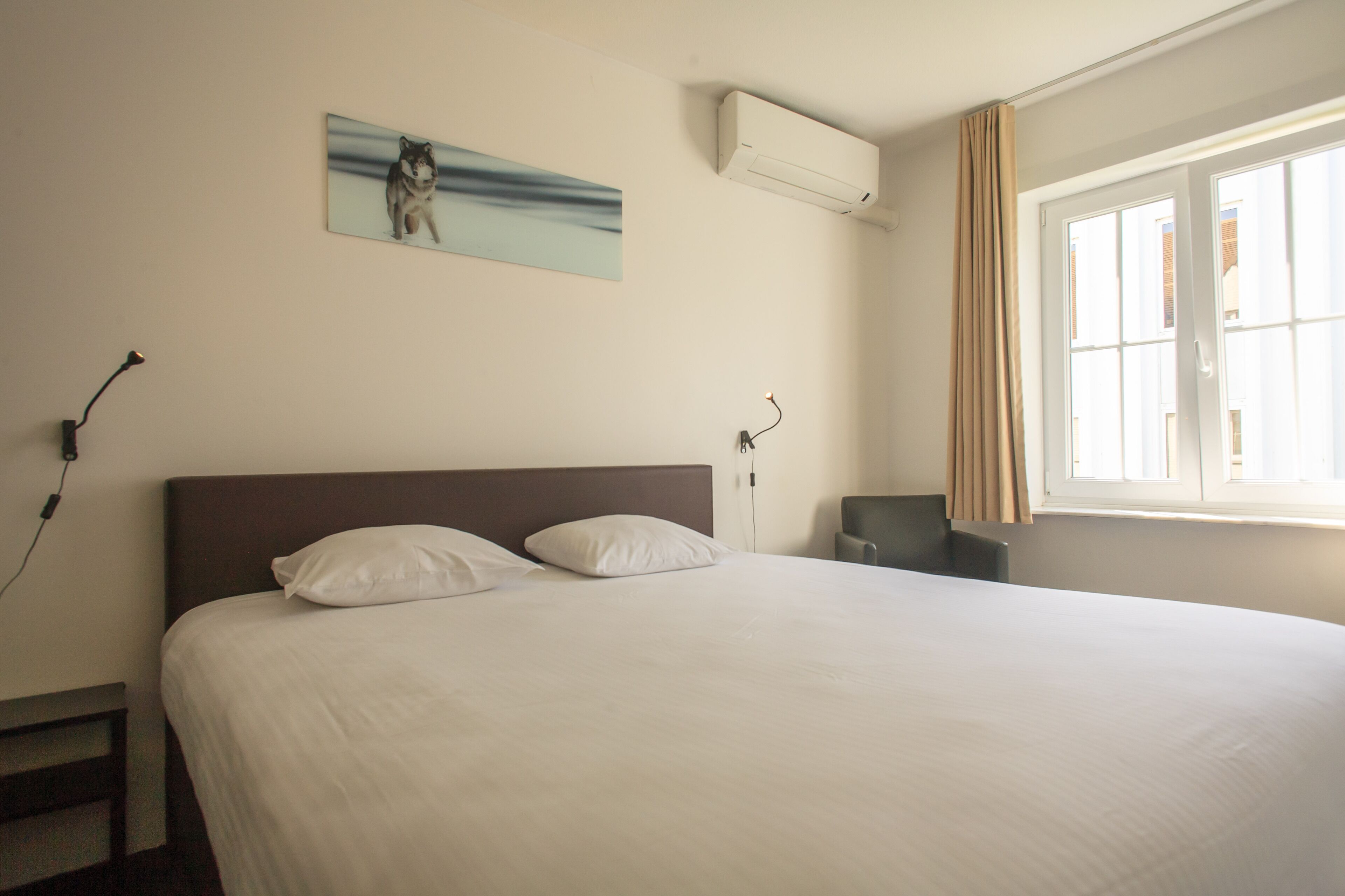 A hotel room with a large bed, a nightstand, a desk, a chair, a window with curtains, and a wall-mounted air conditioner.
