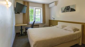 Una habitación de hotel con cama, escritorio con silla, televisor montado en la pared y ventana con cortinas.