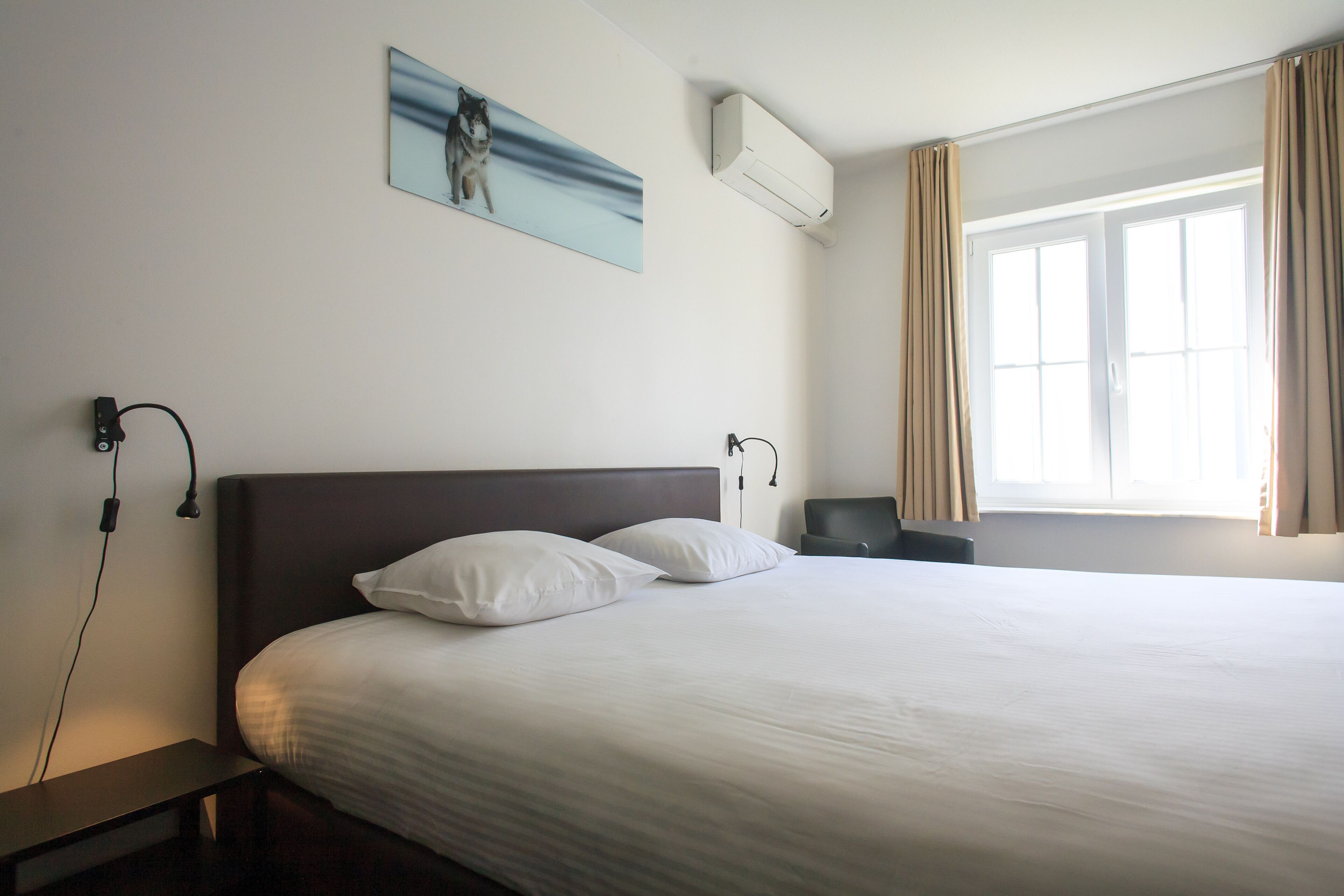 Habitación de hotel con una cama grande, una mesita de noche, un equipo de aire acondicionado, un cuadro en la pared y una ventana con cortinas.