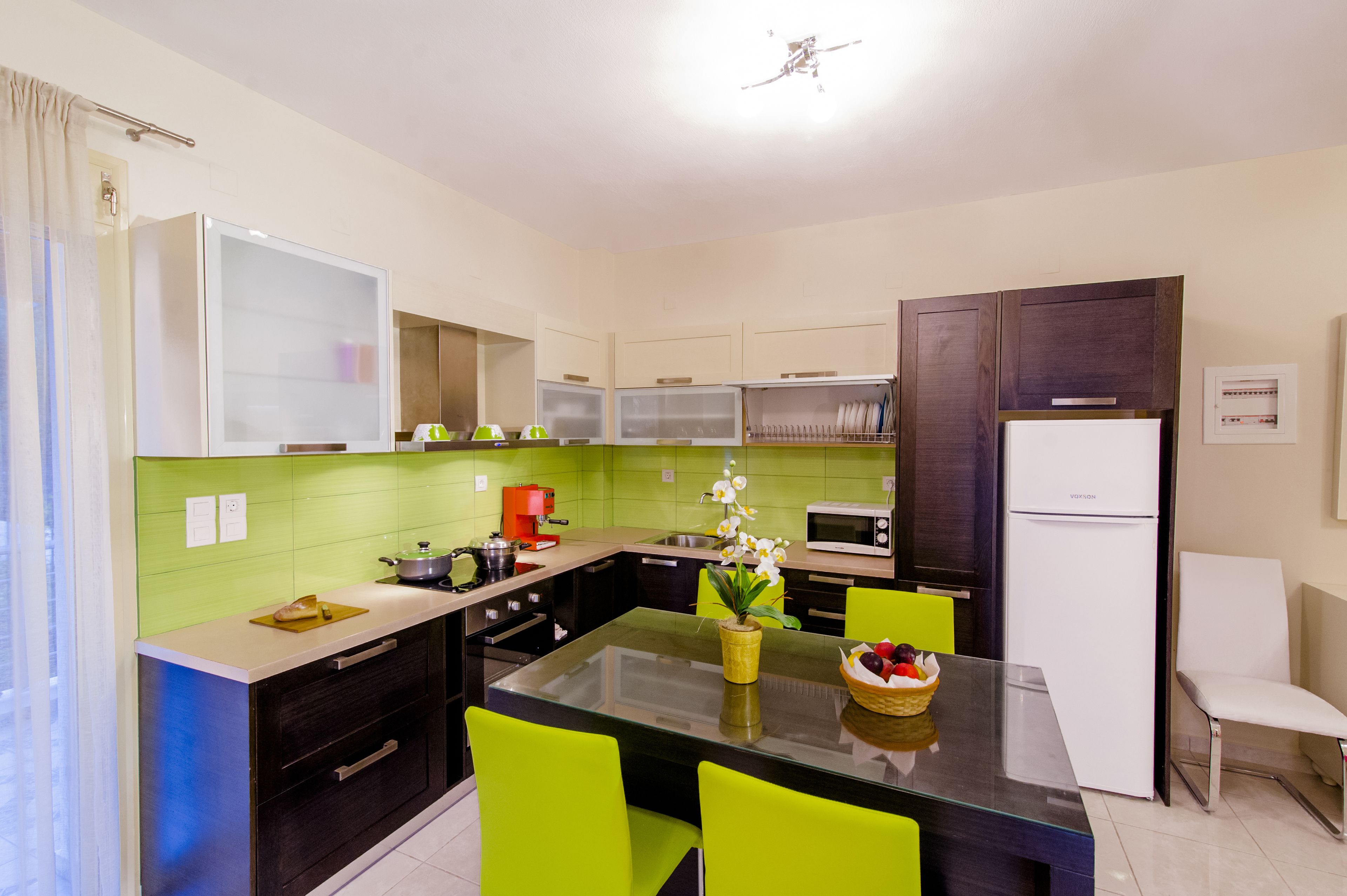 Une cuisine moderne avec des carreaux verts, un îlot central, des chaises d’un vert vif et un réfrigérateur blanc.