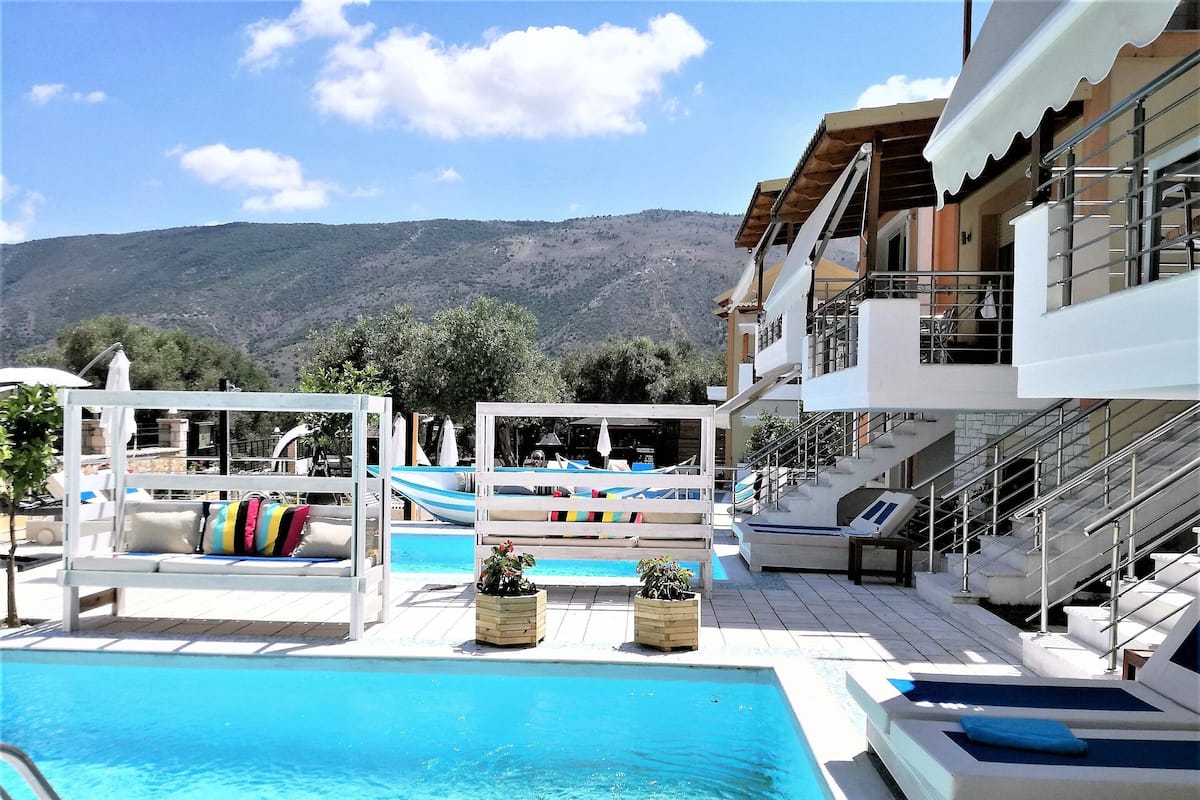 Zona de piscina con sillones reclinables y sombrillas, una escalera y vistas a colinas al fondo.