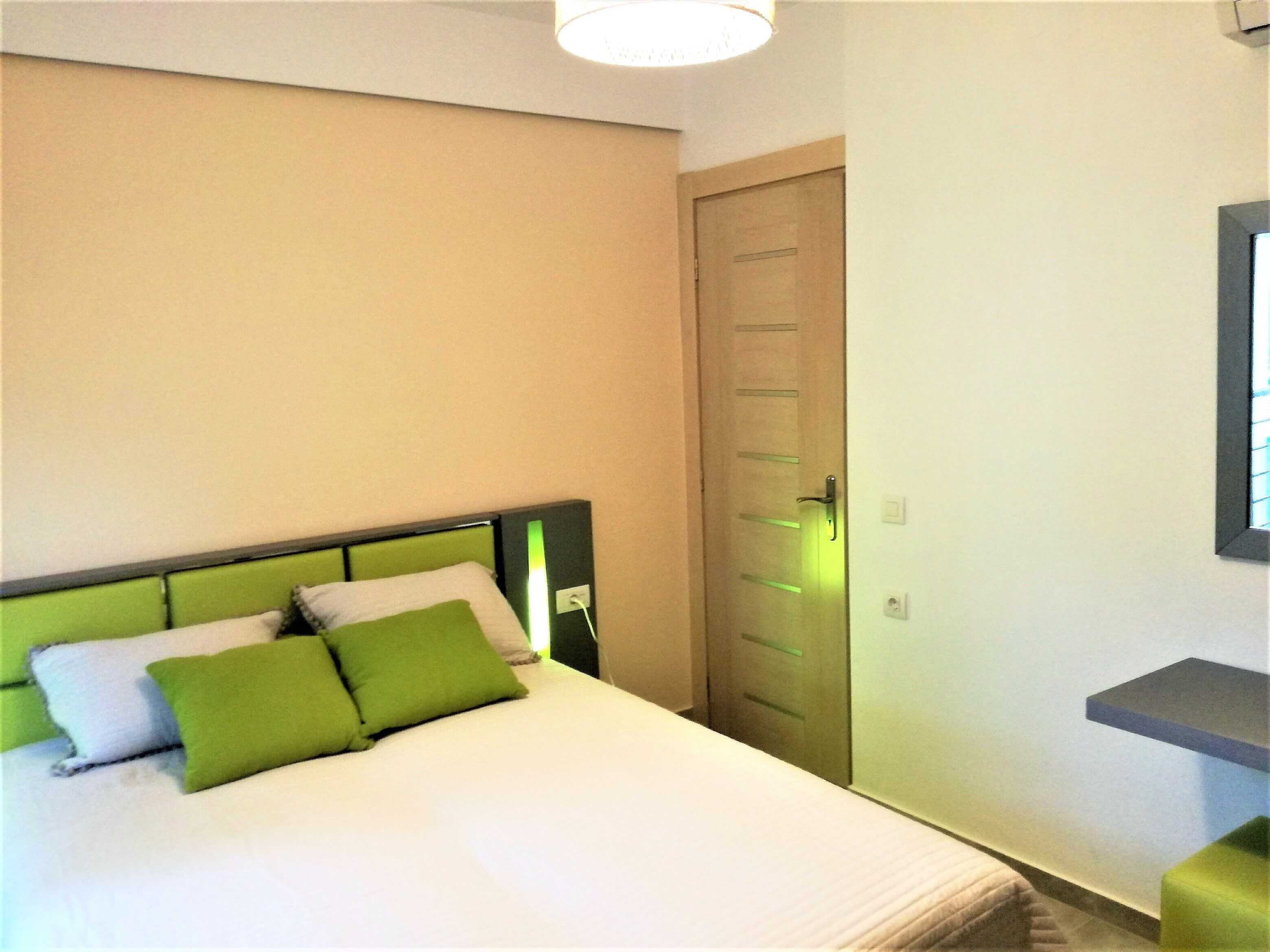 Una habitación de hotel con una cama, cabecera verde, puerta de madera y un estante pequeño.