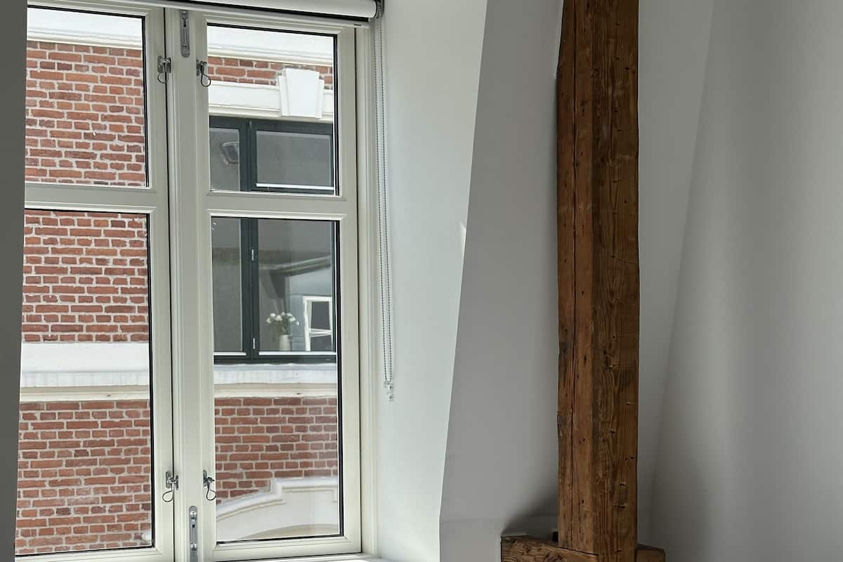 A room with a sloped ceiling, a radiator, a window with a view of a brick building, and a wooden beam.