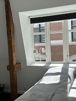 Una habitación con techo inclinado, cama con ropa blanca, ventana con vista a un edificio de ladrillo y estructura de vigas de madera.