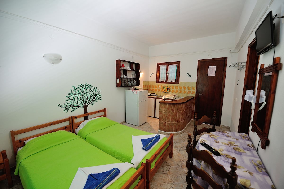 A small, compact room with two single beds, a small kitchenette, a wooden cabinet, and a television mounted on the wall.