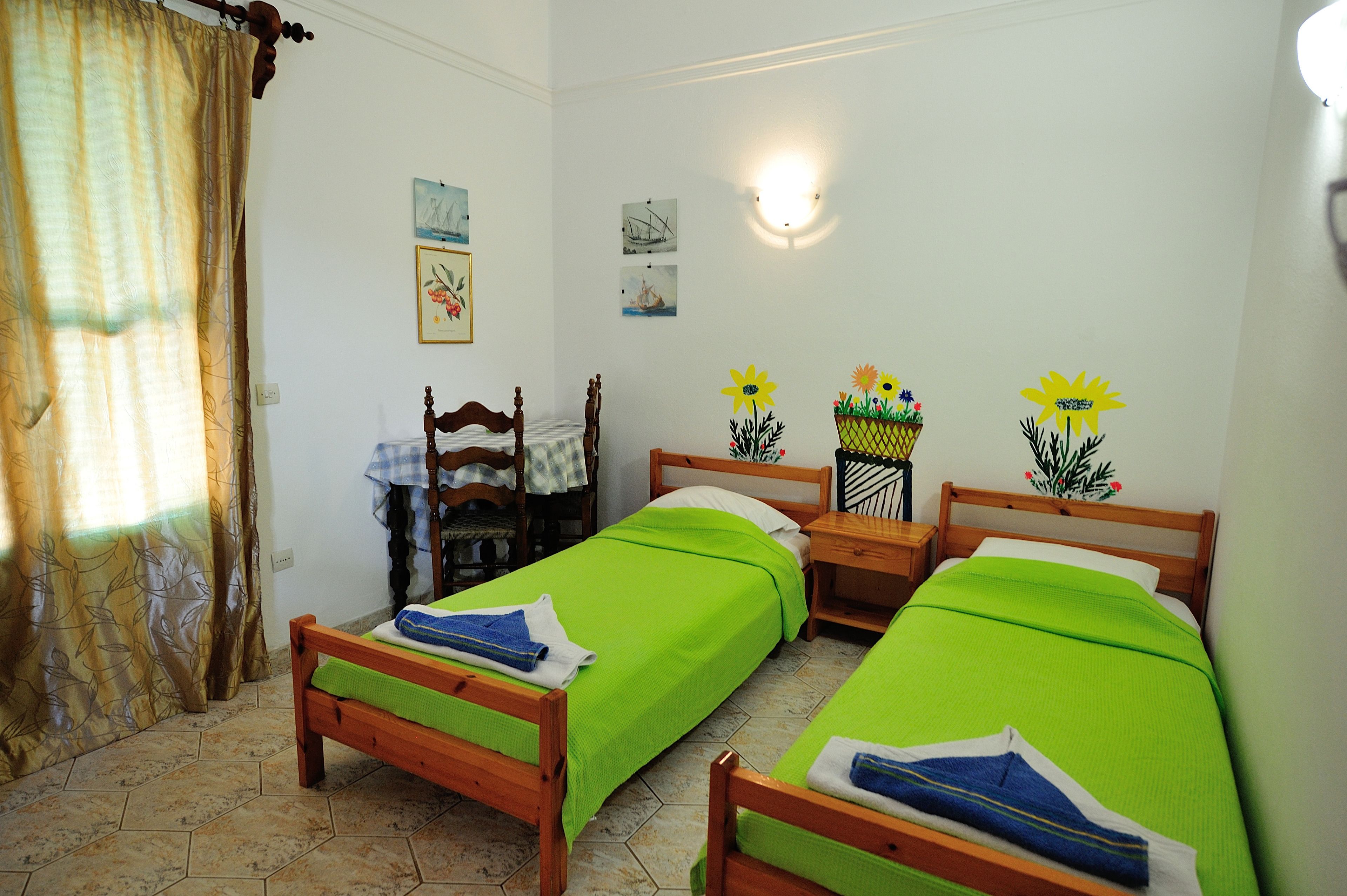 A room with two wooden beds, a wooden nightstand, a wooden chair, a small table with a chair, and a wall-mounted lamp.