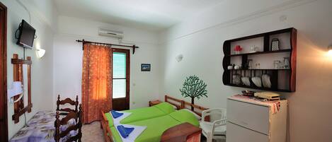 A compact room with a bed, a desk, a chair, a small refrigerator, and a shelf with kitchen items.