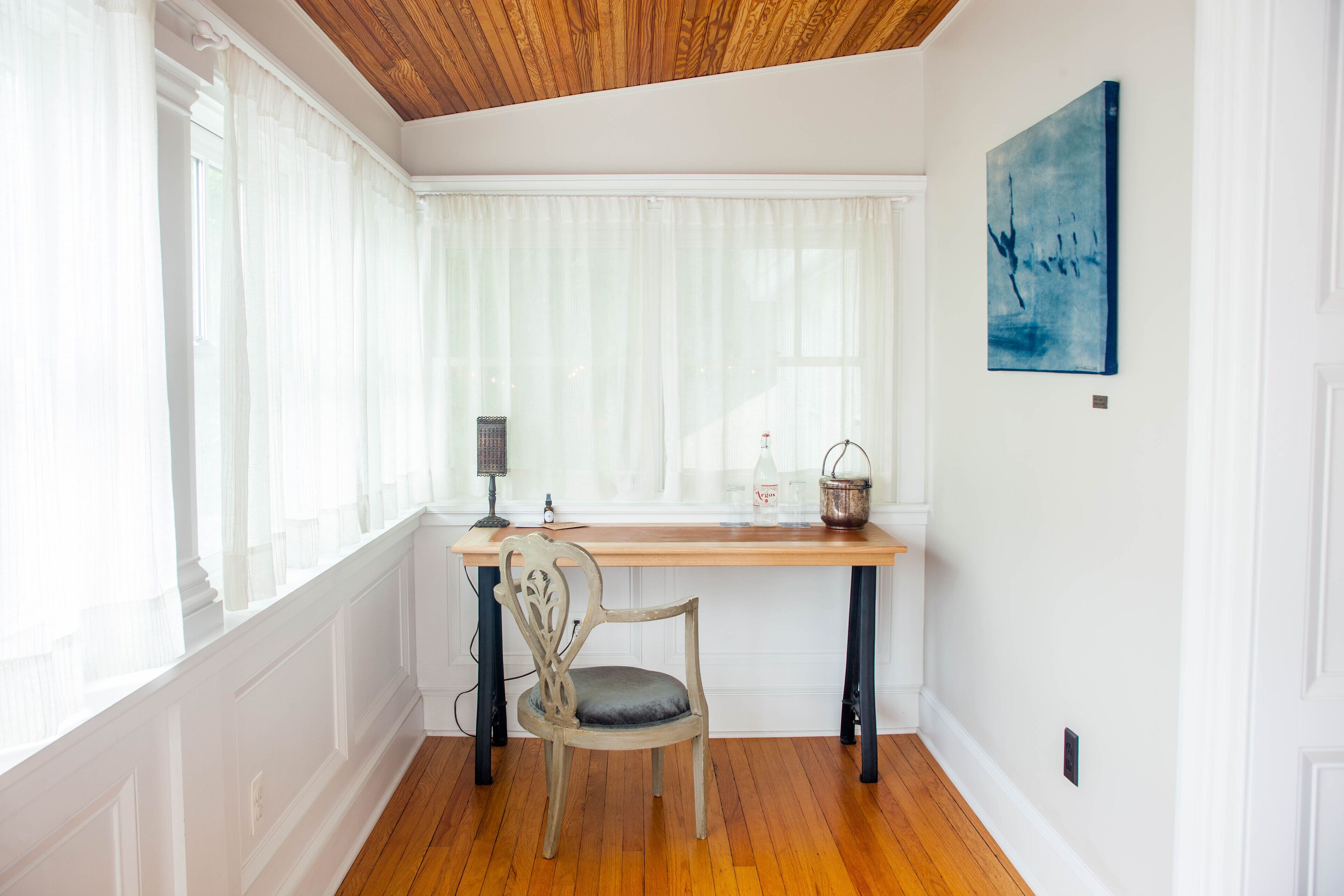 Une pièce avec un bureau en bois, une chaise, une fenêtre avec des rideaux légers, un tableau accroché au mur et un plancher en bois.