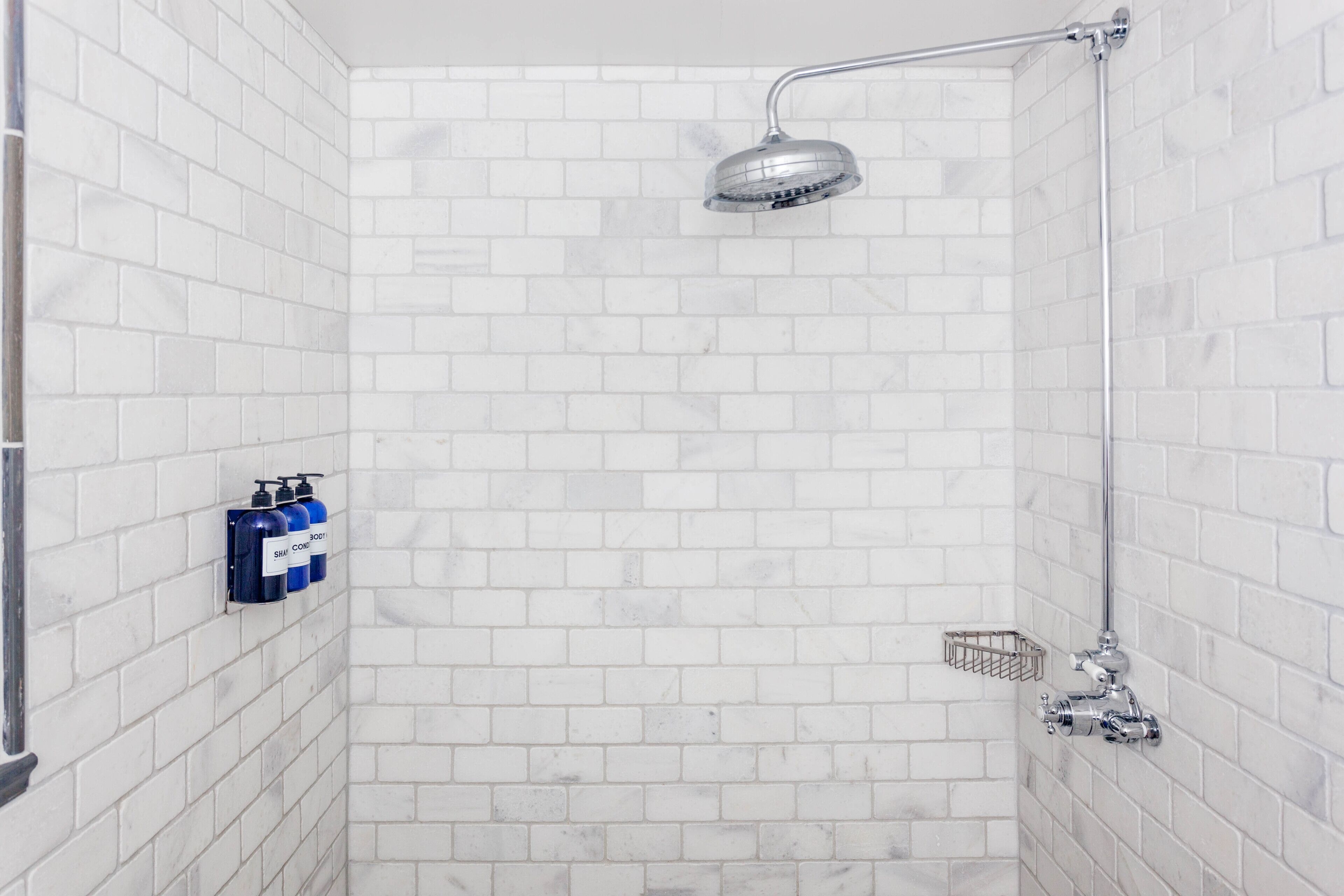 Una ducha con paredes de azulejos blancos, cabezal de ducha cromado y dos botellas de champú y acondicionador.
