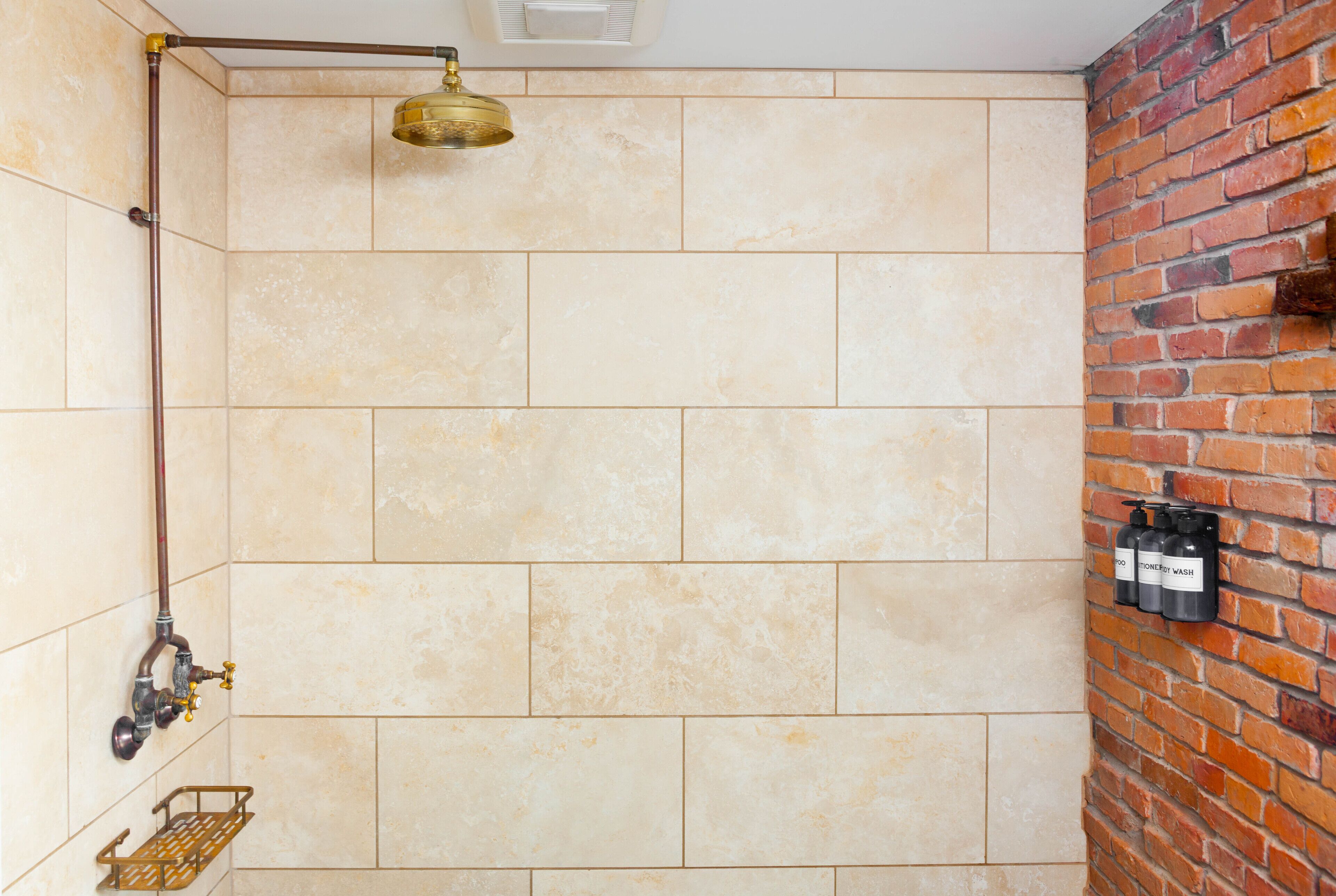 A shower with beige tiles, a brass showerhead, and a brick wall.