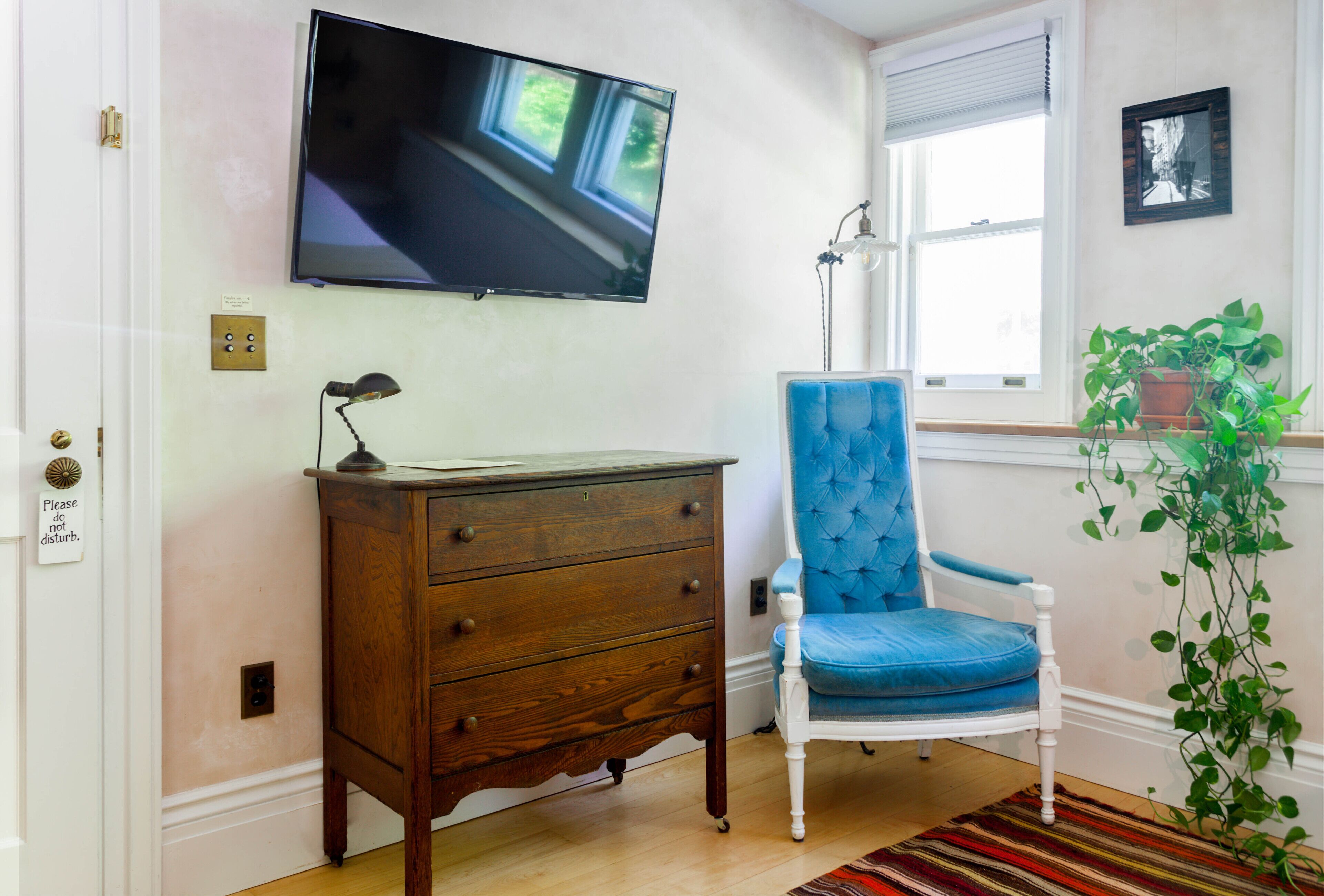 Una habitación con un aparador de madera, un sillón tapizado en azul, un televisor de pantalla plana en la pared, una planta en maceta y una ventana con persianas.